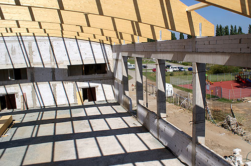 Wnętrze budynku w stanie surowym otwartym z widoczną drewnianą konstrukcją dachu, betonowymi słupami i ścianami z bloczków; w tle plac zabaw i parking.